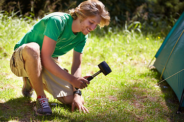 Image showing Man, smile and tent with hammer at campsite for setting up in grass of nature, park and environment. Male person, working and happy with job, rope and outdoor for adventure in summer holiday