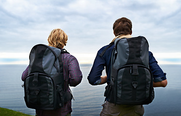 Image showing Couple, backpack and hiking by ocean for travel, sightseeing or outdoor journey in nature. Rear view of man, woman or hikers carrying bag on back for trekking, fitness or adventure with cloudy sky