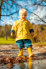 Image showing Sun always shines after the rain. Small bond infant boy wearing yellow rubber boots and yellow waterproof raincoat walking in puddles in city park on sunny rainy day.