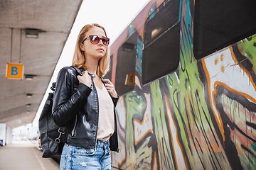 Image showing Young blond woman in jeans, shirt and leather jacket wearing bag and sunglass, embarking modern speed train on train station platform. Travel and transportation.