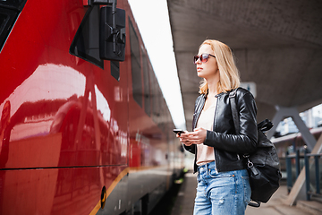 Image showing Young blond woman in jeans, shirt and leather jacket wearing bag and sunglass, embarking red modern speed train on train station platform. Travel and transportation.