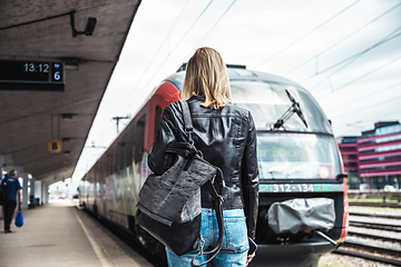 Image showing Young blond woman in jeans, shirt and leather jacket wearing bag and sunglass, embarking red modern speed train on train station platform. Travel and transportation.