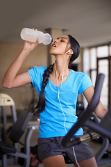 Image showing Woman, drinking water and bike in gym, cycling and stationary machine for workout at club. Female person, earphones and hydrate for strength training, music and equipment for fitness and body health