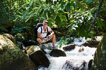 Image showing Man, hiking and river in tropical rainforest with smile for adventure, journey and rocks with backpack. Person, outdoor and bag by water, earth and trees in jungle on holiday with freedom in Colombia