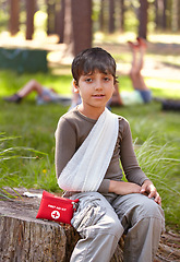 Image showing Child, portrait and first aid for hurt arm with sling on bruise for medical treatment outdoors on tree trunk. Kid, emergency kit and bandage for muscle sprain, injury and camp accident in nature.