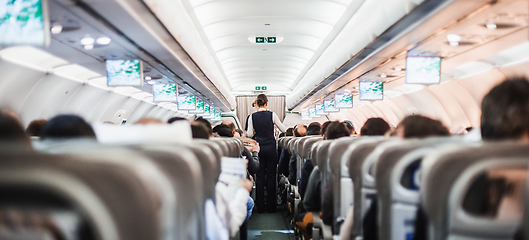 Image showing Interior of airplane with passengers on seats and stewardess in uniform walking the aisle, serving people. Commercial economy flight service concept.