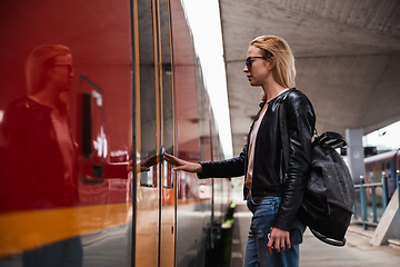 Image showing Young blond woman in jeans, shirt and leather jacket wearing bag and sunglass, presses door button of modern speed train to embark on train station platform. Travel and transportation.