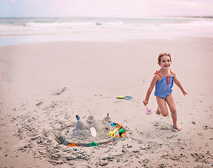 Image showing Portrait, beach and little girl running with sandcastle outdoor by sea or ocean for carefree fun. Nature, water and energy with happy young child playing on sand at coast for holiday or vacation
