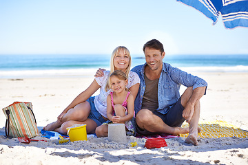 Image showing Parents, girl and sand castle in portrait by ocean, blanket and umbrella with hug on holiday in summer. Father, mother and daughter with picnic at beach for vacation in nature with love in sunshine