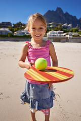 Image showing Young girl, smile and tennis ball in beach with bat, playing and active outdoor. Child, motor skills and growth for development in ocean or sea with mountain view, sand and enjoying in summer