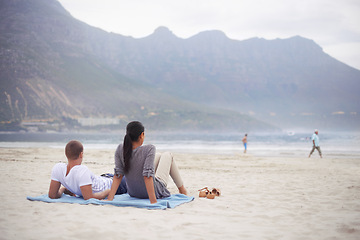 Image showing Couple, beach and vacation on blanket, picnic or relaxing on seashore by mountains. Romantic, honeymoon and love between happy husband and wife, married and date on ocean coast enjoying sunshine