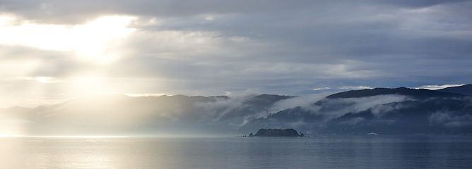 Image showing Ocean, sunset and cloud on mountains in morning, cold and tropical island for vacation in nature. Blue sky, clouds and sunlight on water on beach, calm and winter weather on sustainable environment