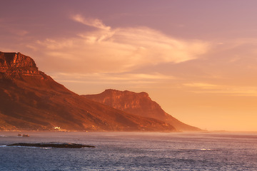 Image showing Ocean, sunset and clouds on mountains by blue sky and outdoor travel for vacation in nature. Landscape, sea and sunlight on false bay with calm and seal island in cape town for tourist destination
