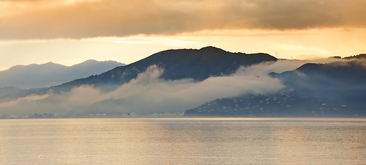 Image showing Sea, sunset and fog in clouds with mountains, water and winter season in evening. New Zealand, travel and destination for holiday in island, landscape and environment for vacation with scenery