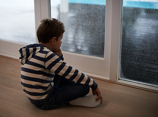 Image showing Rain, window and sad boy child on a floor unhappy, upset or annoyed by bad weather in his home. Glass, depression and kid in a living room frustrated by storm, cold or unexpected winter disaster