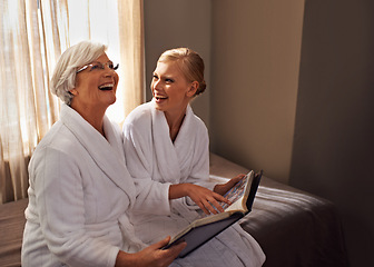 Image showing Grandmother, woman and laughing at photo album, home and happy for bonding and looking at memories. Senior lady, daughter and moment for self care, robe and bedroom for comfort and hospitality