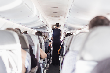 Image showing Interior of airplane with passengers on seats and stewardess in uniform walking the aisle, serving people. Commercial economy flight service concept.