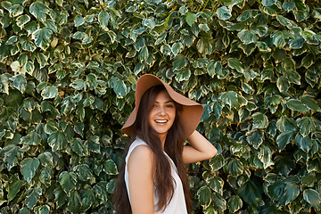 Image showing Woman, portrait and happiness by hedge outdoor with travel, summer holiday and casual fashion with hat. Girl, face and smile at leaf fence in garden for relax, wellness and confidence in Amsterdam