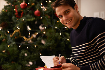 Image showing Portrait, Christmas tree and man writing on present in home for preparation of holiday event of tradition. Box, gift card and smile with young person in apartment for December celebration or vacation