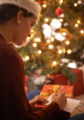 Image showing Woman, writing letter and Christmas in home by tree, present and santa hat with message for festive wish. Girl, person and paper with notes, gift and package with kindness, celebration and xmas card