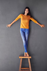 Image showing Woman, clothes and stand on chair in portrait, smile and fashion on wall background. Female person, cool and happy lady on stool, confident and apparel in living room at home in house for trends