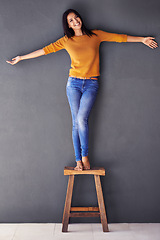 Image showing Woman, confident and stool in fashion portrait, smile and style on wall background. Female person, clothing and happy lady on chair, proud and excited in living room at home in house with balance