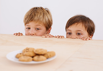 Image showing Children, table and peeking at cookies for curiosity, observation and sneaking with glance. Anticipation, wonder and kids by desk with biscuits for hunger, hiding and playful brothers at home