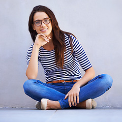 Image showing Portrait, fashion and smile of woman on floor with wall background for casual or trendy style. Happy, model and relax with confident young person legs crossed in glasses for casual clothing outfit