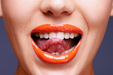 Image showing Beauty, lipstick and woman with tongue out, makeup and closeup of shiny cosmetic product with skin. Lick, flirting and orange lip gloss for aesthetic in studio with cosmetology on blue background