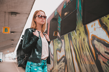 Image showing Young blond woman in jeans, shirt and leather jacket wearing bag and sunglass, embarking modern speed train on train station platform. Travel and transportation.