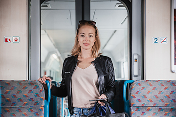 Image showing Young blond woman in jeans, shirt and leather jacket holding her smart phone and purse while riding modern speed train arriving to final train station stop. Travel and transportation.