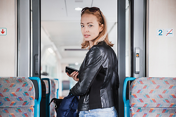 Image showing Young blond woman in jeans, shirt and leather jacket holding her smart phone and purse while riding modern speed train arriving to final train station stop. Travel and transportation.