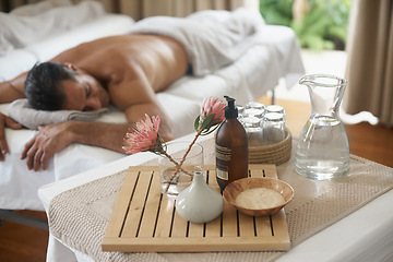 Image showing Salt, flowers and man in spa to relax on bed or break with luxury pamper treatment tools on table. Protea, facial oil or person at resort and salon for natural healing benefits or massage with water