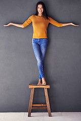 Image showing Woman, fashion and stand on chair in portrait, smile and balance on wall background. Female person, modern clothing and happy lady on stool, barefoot and confidence in living room at home in house