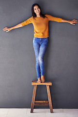 Image showing Woman, confident and smile on chair in portrait, stand and fashion on wall background. Female person, studio and happy lady on stool, barefoot and balance in living room at home with modern clothing