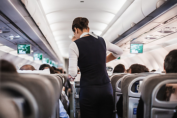 Image showing Interior of airplane with passengers on seats and stewardess in uniform walking the aisle, serving people. Commercial economy flight service concept.