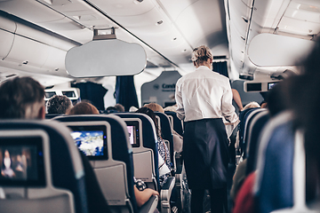 Image showing Interior of airplane with passengers on seats and stewardess in uniform walking the aisle, serving people. Commercial economy flight service concept.