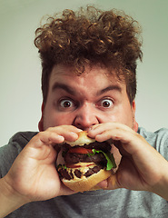 Image showing Takeaway, burger and food for man in studio with meal for big eater, plus size or greed. Young person with meat in mouth for hunger, silly and bite indoor for delicious, taste and sauce for portrait