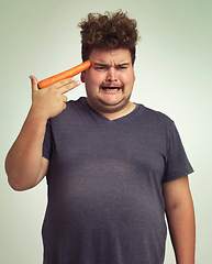 Image showing Depression, carrot and hand gun with plus size man in studio on gray background for diet, health or nutrition. Sad, unhappy and vegetable to head of young person in conflict with vegetarian food