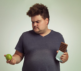 Image showing Man, apple and chocolate in studio with choice for diet, healthy food and nutrition for wellness. Male person, plus size and thinking with decision for fruit, sweet dessert and snack for lifestyle