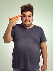 Image showing Portrait, vegetable and hand gun with plus size man in studio on gray background for diet, health or nutrition. Anger, depression and carrot to head of young person in conflict with vegetarian food