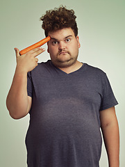 Image showing Portrait, carrot and hand gun with plus size man in studio on gray background for diet, health or nutrition. Depression, sad and vegetable to head of young person in conflict with vegetarian food