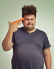 Image showing Depression, vegetable and hand gun with plus size man in studio on gray background for diet, health or nutrition. Sad, unhappy and carrot to head of young person in conflict with vegetarian food