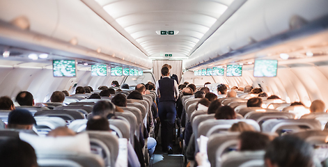 Image showing Interior of airplane with passengers on seats and stewardess in uniform walking the aisle, serving people. Commercial economy flight service concept.