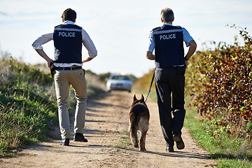 Image showing Policeman, dog and walk in field for crime scene or robbery with car for search, safety and law enforcement. Detective, investigation and uniform in outdoor working at countryside with gravel road