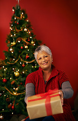 Image showing Christmas, senior and happy woman with gift in portrait at studio for holiday celebration by tree isolated on a red background. Xmas, giving and person with present or offer box for festive party