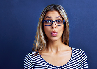 Image showing Funny, face and woman with glasses in studio for tongue out, silly or humor on blue background. Need, portrait and female model with emoji expression, gesture or geek, aesthetic or quirky personality