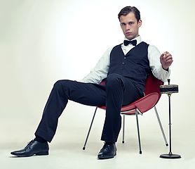 Image showing Fashion, tuxedo and portrait of man in studio with red chair and cigarette for trendy, elegant and fancy outfit. Serious, handsome and person with bowtie and suit for classy style by white background