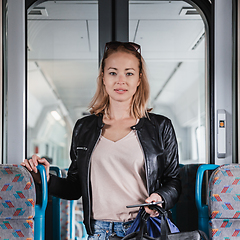 Image showing Young blond woman in jeans, shirt and leather jacket holding her smart phone and purse while riding modern speed train arriving to final train station stop. Travel and transportation.