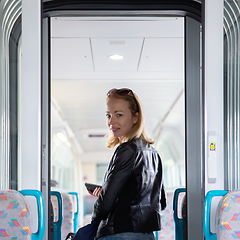 Image showing Young blond woman in jeans, shirt and leather jacket holding her smart phone and purse while riding modern speed train arriving to final train station stop. Travel and transportation.